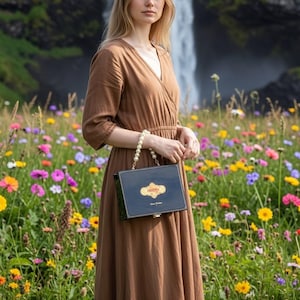 May include: A woman in a brown dress holds a navy blue handbag with a pearl handle. The handbag has a gold emblem and text. The background features a waterfall and a field of colorful wildflowers.