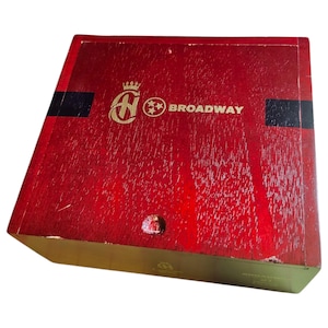 Peut inclure: Une boîte en bois rouge avec un couvercle à charnière. La boîte présente un emblème doré et le mot "BROADWAY" en lettres dorées. Des rayures noires sont sur le dessus. La boîte est inclinée, révélant son côté.