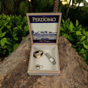 May include: A wooden box with a hinged lid, featuring the text "PERDOMO LOT 23" and a blue and white illustration of a landscape. The box is open, revealing a white interior with two watches and two rings.