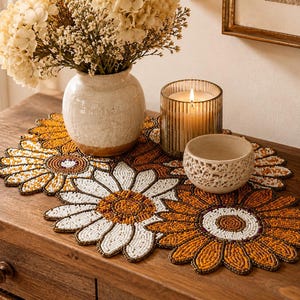 May include: A beaded table runner featuring a floral pattern of large daisy-like flowers in shades of white, orange, and brown. The runner is composed of small, uniform beads arranged in circular and petal shapes, creating a textured surface.