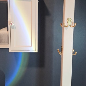 Può includere: Un mobiletto bianco a parete con pomello floreale ed effetto luce arcobaleno. Un attaccapanni bianco con ganci dorati è nelle vicinanze. Due piante in vaso sono su una mensola sopra il mobile. Il muro è dipinto di blu scuro.