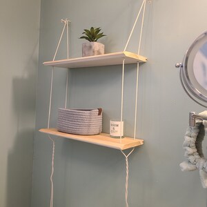 May include: Two wooden shelves hanging from the ceiling with white rope. The top shelf has a small potted succulent plant. The bottom shelf has a gray woven basket and a white candle with the text "Warm & Cozy".