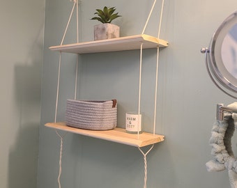 May include: Two wooden shelves hanging from the ceiling with white rope. The top shelf has a small potted succulent plant. The bottom shelf has a gray woven basket and a white candle with the text "Warm & Cozy".
