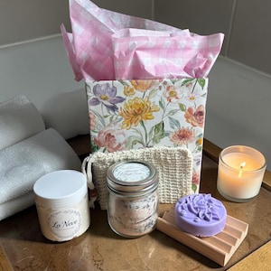 May include: A gift set with a floral-patterned box, pink gingham tissue paper, and various bath products. Includes a jar of bath salts, a lavender soap bar on a wooden holder, a candle, and a jar of cream labeled "La Neve".