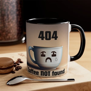 May include: A white coffee mug with a black interior and handle. The mug features a sad coffee cup graphic with the text "404 coffee NOT found". The mug is on a wooden surface with a spoon and a macaron.