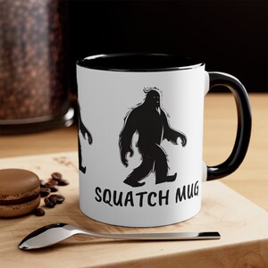 Könnte beinhalten: Weiße Keramiktasse mit schwarzem Rand und Henkel. Die Tasse zeigt eine schwarze Silhouette eines Bigfoots, der geht, und den Text "Squatch Mug".