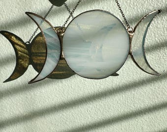 May include: A triple moon pendant made of stained glass with a white iridescent finish. The pendant is hanging from a chain and is in focus against a light green background.