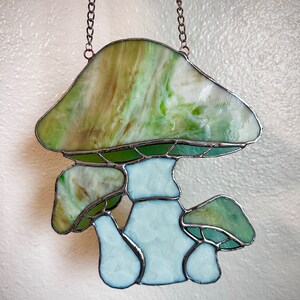 Op de afbeelding: Een gebrandschilderd glazen zonnenvanger in de vorm van een paddenstoel met een groen-wit swirl-ontwerp. De paddenstoel heeft een grote hoed en een steel met twee kleinere paddenstoelen die eruit groeien.