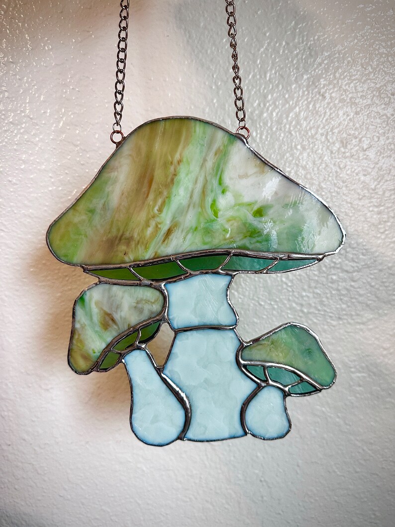 Op de afbeelding: Een gebrandschilderd glazen zonnenvanger in de vorm van een paddenstoel met een groen-wit swirl-ontwerp. De paddenstoel heeft een grote hoed en een steel met twee kleinere paddenstoelen die eruit groeien.