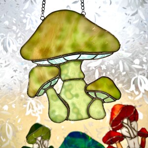 Op de afbeelding: Een gebrandschilderd glazen zonvanger in de vorm van een paddenstoel met een groen en wit ontwerp. De zonvanger hangt aan een ketting en heeft drie paddenstoelen met verschillende hoedgroottes.