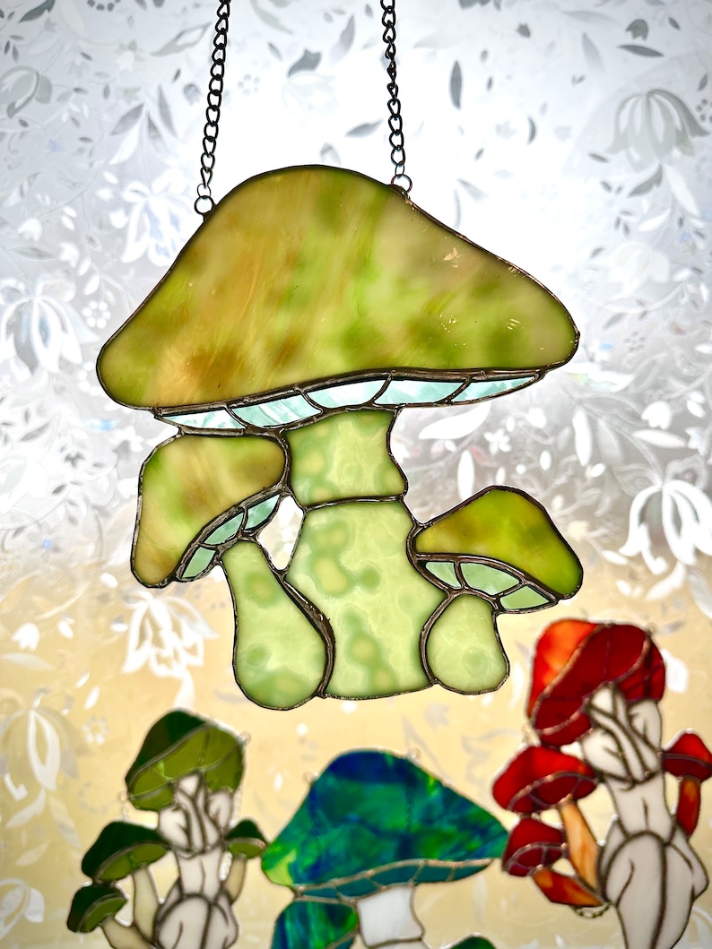 Op de afbeelding: Een gebrandschilderd glazen zonvanger in de vorm van een paddenstoel met een groen en wit ontwerp. De zonvanger hangt aan een ketting en heeft drie paddenstoelen met verschillende hoedgroottes.