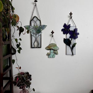 Op de afbeelding: Drie gebrandschilderde wanddecoraties met een groene vlinder, een paarse bloem en een groene paddenstoel. De decoraties zijn allemaal rechthoekig en hebben een witte achtergrond.