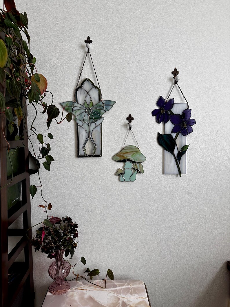 Op de afbeelding: Drie gebrandschilderde wanddecoraties met een groene vlinder, een paarse bloem en een groene paddenstoel. De decoraties zijn allemaal rechthoekig en hebben een witte achtergrond.