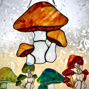 Op de afbeelding: Een gebrandschilderd glazen zonnenvanger in de vorm van een paddenstoel met een rode en oranje dop en een witte steel. De paddenstoel hangt aan een ketting en er zijn andere gebrandschilderde glazen paddenstoelen op de achtergrond.