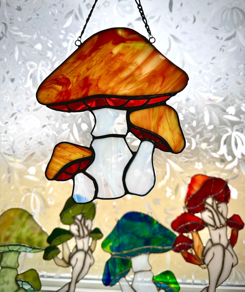 Op de afbeelding: Een gebrandschilderd glazen zonnenvanger in de vorm van een paddenstoel met een rode en oranje dop en een witte steel. De paddenstoel hangt aan een ketting en er zijn andere gebrandschilderde glazen paddenstoelen op de achtergrond.