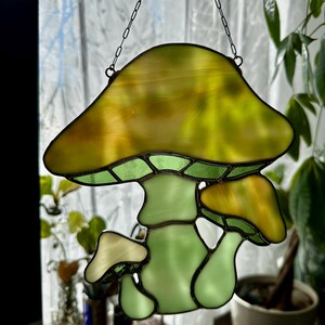 Op de afbeelding: Een hangende paddenstoel-versiering van gekleurd glas met een groene steel en een gele hoed. De versiering hangt aan een ketting.
