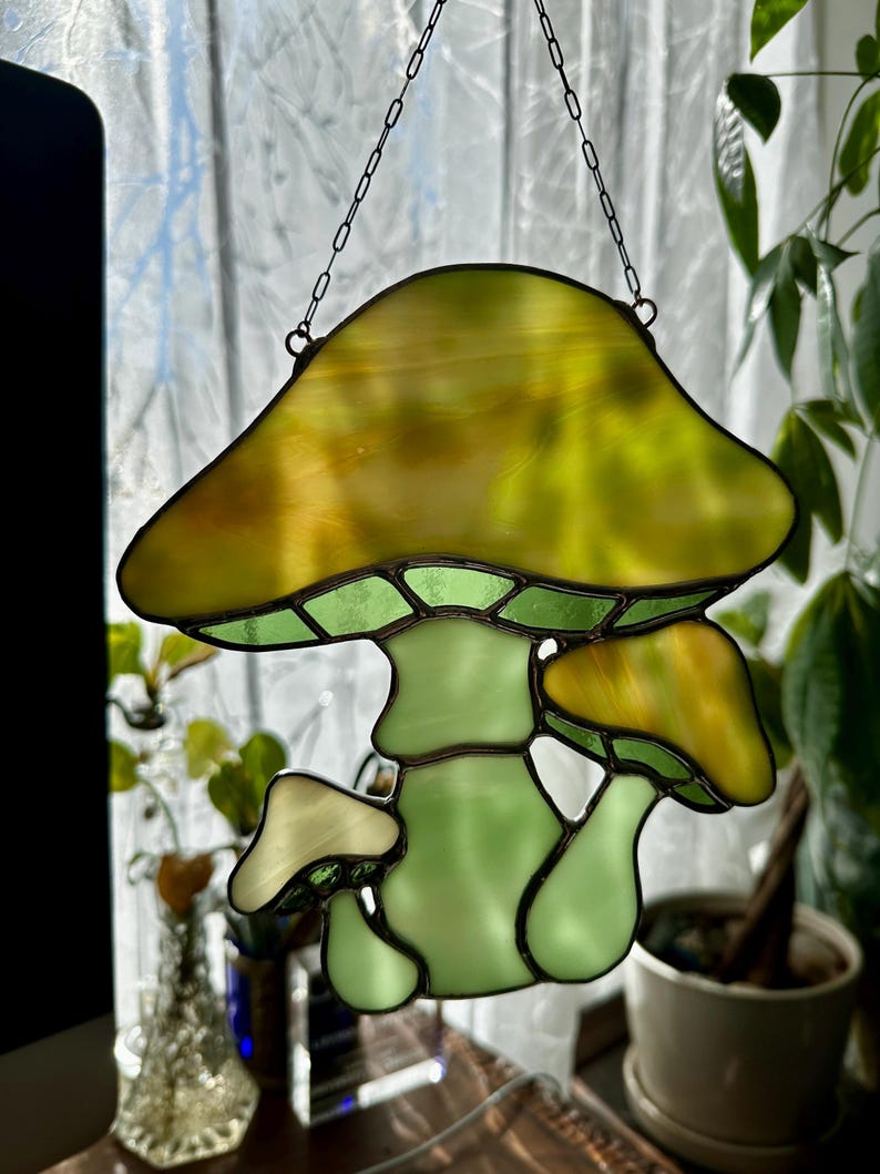 Op de afbeelding: Een hangende paddenstoel-versiering van gekleurd glas met een groene steel en een gele hoed. De versiering hangt aan een ketting.