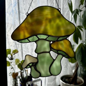 Op de afbeelding: Een gebrandschilderd glazen zonenvanger in de vorm van een paddenstoel met een gele bovenkant en een groene steel. De zonenvanger hangt aan een ketting en heeft drie paddenstoelen.