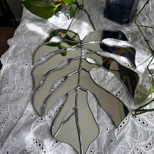 Op de afbeelding: Een zilverkleurige spiegel in de vorm van een monstera-blad. De spiegel hangt aan een ketting en reflecteert de omgeving.