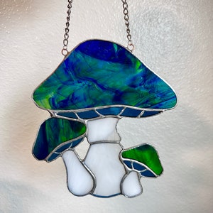 Op de afbeelding: Een gebrandschilderd paddenstoel zonenvanger met een blauwe en groene bovenkant en een witte steel. De zonenvanger hangt aan een zilveren ketting.
