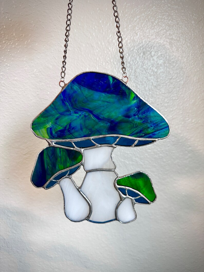 Op de afbeelding: Een gebrandschilderd paddenstoel zonenvanger met een blauwe en groene bovenkant en een witte steel. De zonenvanger hangt aan een zilveren ketting.