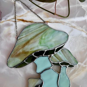 Op de afbeelding: Een gebrandschilderd glazen paddenstoel zonenvanger met een groene en bruine bovenkant en een lichtblauwe onderkant. De zonenvanger hangt aan een ketting.