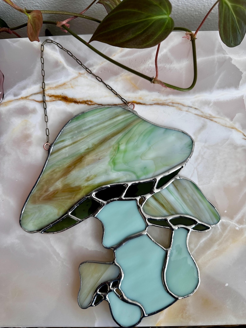 Op de afbeelding: Een gebrandschilderd glazen paddenstoel zonenvanger met een groene en bruine bovenkant en een lichtblauwe onderkant. De zonenvanger hangt aan een ketting.