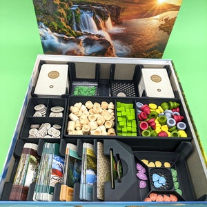 May include: A black and gray organizer insert for the board game "Earth" by Conor McGoey. The insert has compartments for cards, tokens, and other game components. The insert is shown in an open box with a scenic image of a waterfall and mountains.