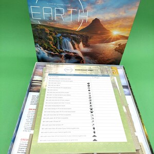 May include: A board game box with a green background. The box is open and shows the game's achievement sheet, which lists various conditions for earning points. The box lid features a scenic image of a mountain range with a waterfall and a sunset.