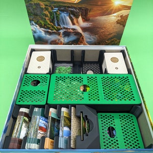 May include: An open box game insert with green and black compartments for organizing game components. The box lid features a landscape image of a waterfall and a mountain range with the text "EARTH" and "PRESENTED BY CONOR MCGREEY".