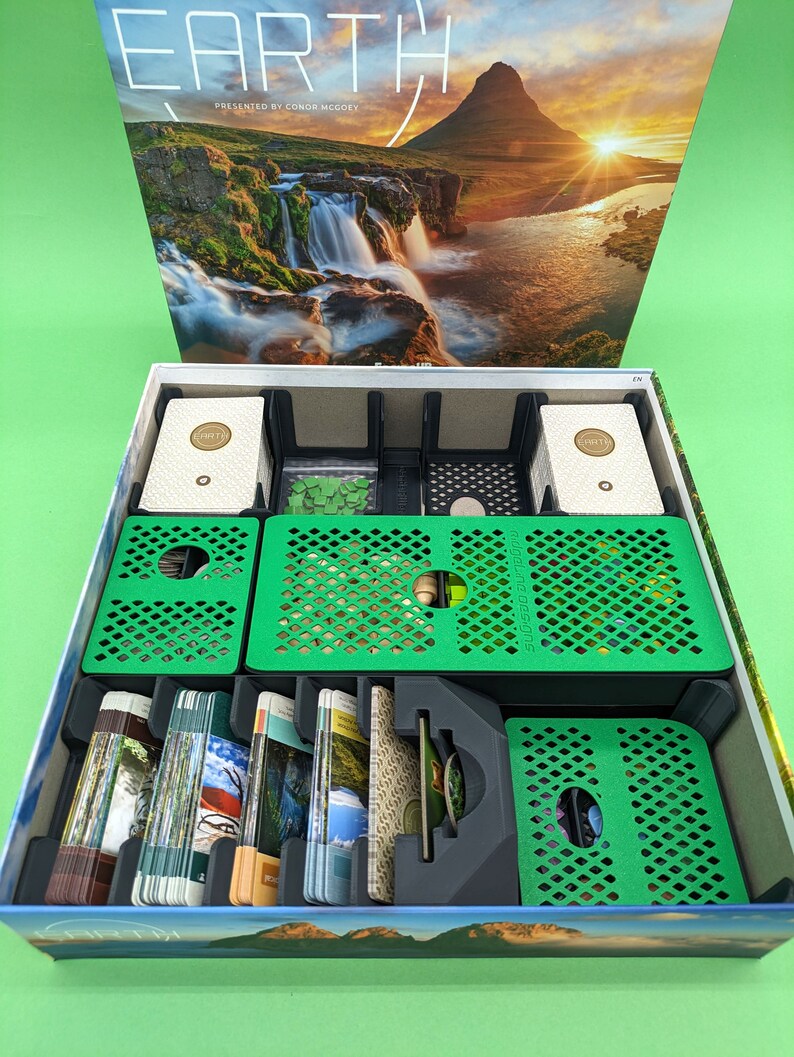 May include: An open box game insert with green and black compartments for organizing game components. The box lid features a landscape image of a waterfall and a mountain range with the text "EARTH" and "PRESENTED BY CONOR MCGREEY".