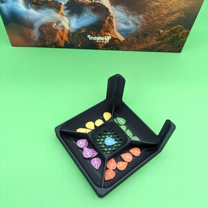 May include: Black game board with a grid in the center and four compartments around the grid. Each compartment has a different color of leaf-shaped game pieces. The game board is on a green surface. "InsideUp Games" is printed on the board.