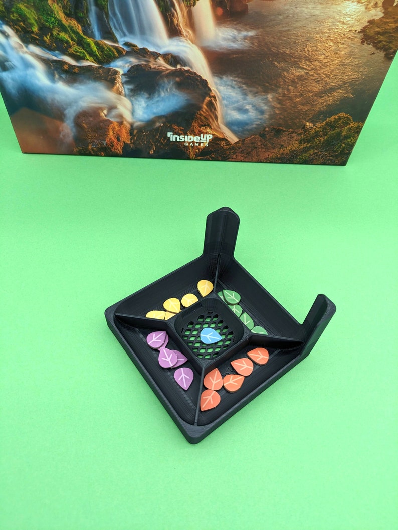 May include: Black game board with a grid in the center and four compartments around the grid. Each compartment has a different color of leaf-shaped game pieces. The game board is on a green surface. "InsideUp Games" is printed on the board.