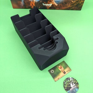May include: Black plastic card holder with multiple slots for organizing cards. The holder is sitting on a green surface. There are two cards and a round token with nature scenes on them.