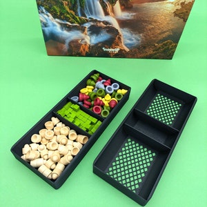 May include: Two black plastic trays with compartments for storing game pieces. The trays are filled with various colored game pieces, including green, red, yellow, and wooden pieces. The trays are on a green surface.