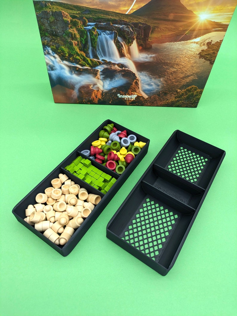 May include: Two black plastic trays with compartments for storing game pieces. The trays are filled with various colored game pieces, including green, red, yellow, and wooden pieces. The trays are on a green surface.