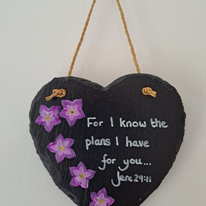 May include: A heart-shaped, black slate plaque with a jute rope hanger. The plaque features hand-painted purple flowers and the text "For I know the plans I have for you... Jere 29:11" in white. A decorative wall hanging.