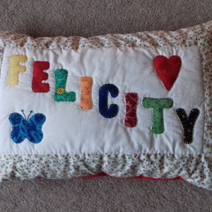 May include: A white pillow with a floral print border. The pillow has the name "FELICITY" spelled out in colorful fabric letters. A red heart and a blue butterfly are also sewn onto the pillow.