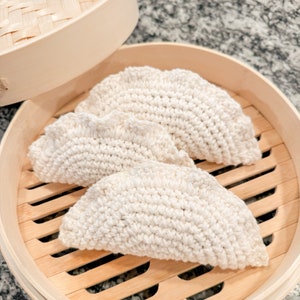 May include: Three crocheted white dumplings in a bamboo steamer. The dumplings are made with a textured yarn and have a slightly ruffled edge.