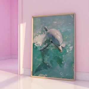 Op de afbeelding: Een ingelijst kunstwerk met een dolfijn die in turquoise water zwemt. De dolfijn is grijs en wit, met een impressionistische stijl. De lijst is lichtbruin en het kunstwerk wordt tentoongesteld aan een roze muur.