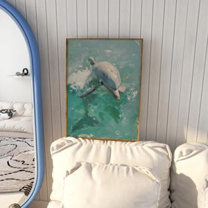 Op de afbeelding: Een ingelijst schilderij van een dolfijn die in turquoise water zwemt. Het kunstwerk heeft een gouden lijst en wordt tentoongesteld tegen een witte muur met verticale panelen. De dolfijn is afgebeeld in tinten grijs en wit.