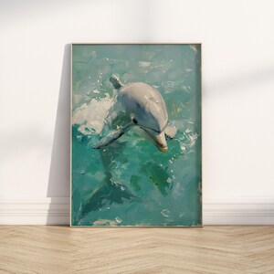 Op de afbeelding: Olieverfschilderij van een dolfijn die zwemt in turquoise water. De dolfijn is lichtgrijs met een witte buik en zijn vorm is vastgelegd met zichtbare penseelstreken. Het kunstwerk is ingelijst in een licht houten frame, leunend tegen een witte muur.