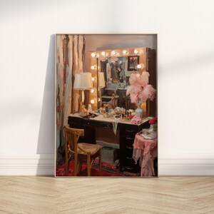May include: An artistic print of a vintage vanity table with a lighted mirror, a wooden chair, and a table with a pink cloth. The vanity is adorned with makeup, jewelry, and a vase of pink feathers. The room has a warm, inviting atmosphere.