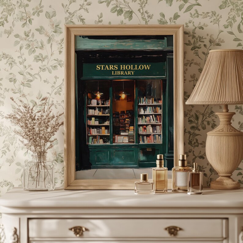 Op de afbeelding: Een ingelijste print van een groene winkelgevel met de woorden "Stars Hollow Library" boven de ingang. De winkelgevel heeft een groot raam met boekenplanken erin. De print staat op een wit oppervlak met een lamp en parfumflesjes ervoor.