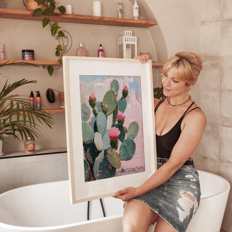 May include: A framed painting of a cactus with pink flowers. The painting is in a light wood frame and is being held by a person in a black tank top and denim skirt.