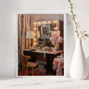 May include: A framed painting depicts a vintage vanity scene. The black vanity is adorned with makeup, jewelry, and a lamp. A large mirror with lights is above the vanity. A vase with flowers is on the right.