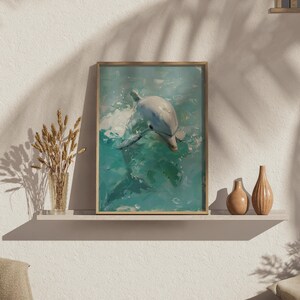 Op de afbeelding: Een ingelijste kunstdruk met een dolfijn die in turquoise water zwemt. Het kunstwerk heeft een textuurrijke, schilderachtige stijl met tinten wit, blauw en groen. De lijst is licht van kleur, op een witte plank.