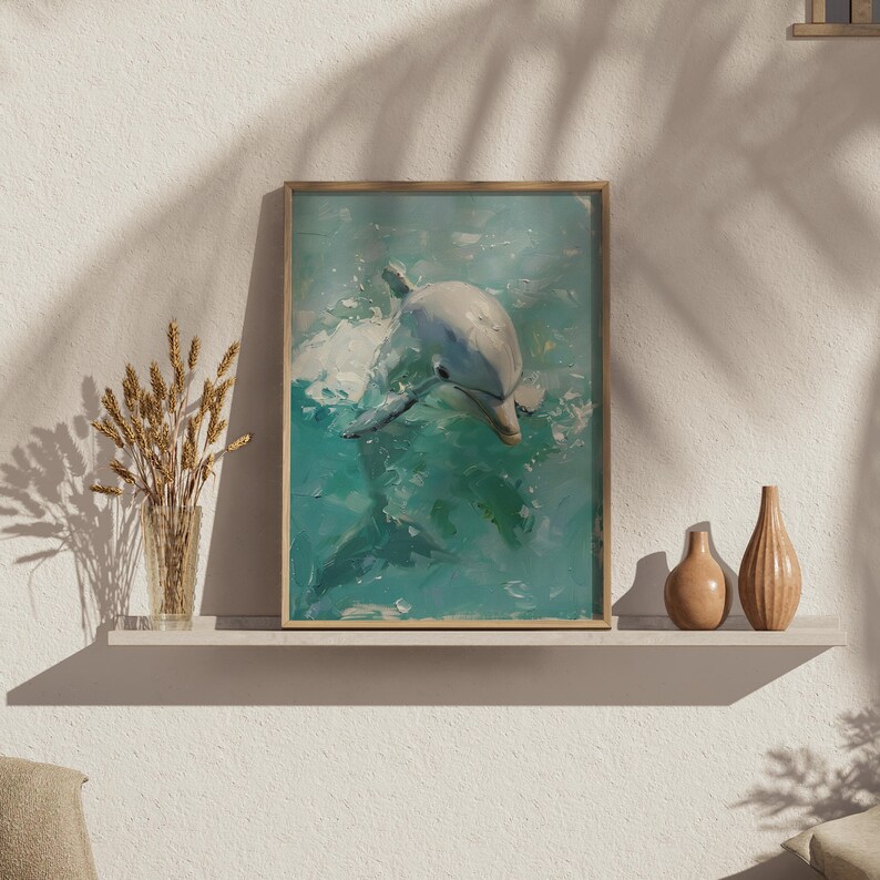 Op de afbeelding: Een ingelijste kunstdruk met een dolfijn die in turquoise water zwemt. Het kunstwerk heeft een textuurrijke, schilderachtige stijl met tinten wit, blauw en groen. De lijst is licht van kleur, op een witte plank.