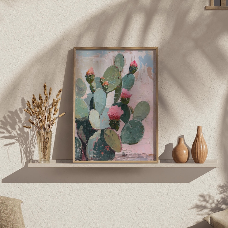 May include: A framed painting of a cactus with pink flowers on a pink background. The cactus is green and has a textured surface. The painting is framed in a light wood frame.