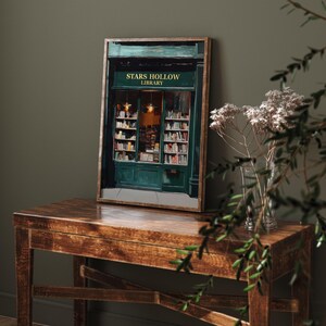 Op de afbeelding: Een ingelijste print van een groene winkelgevel met de woorden "Stars Hollow Library" boven de ingang. De winkelgevel heeft een groot raam met boekenplanken vol boeken. De print staat op een houten tafel met een vaas met witte bloemen.